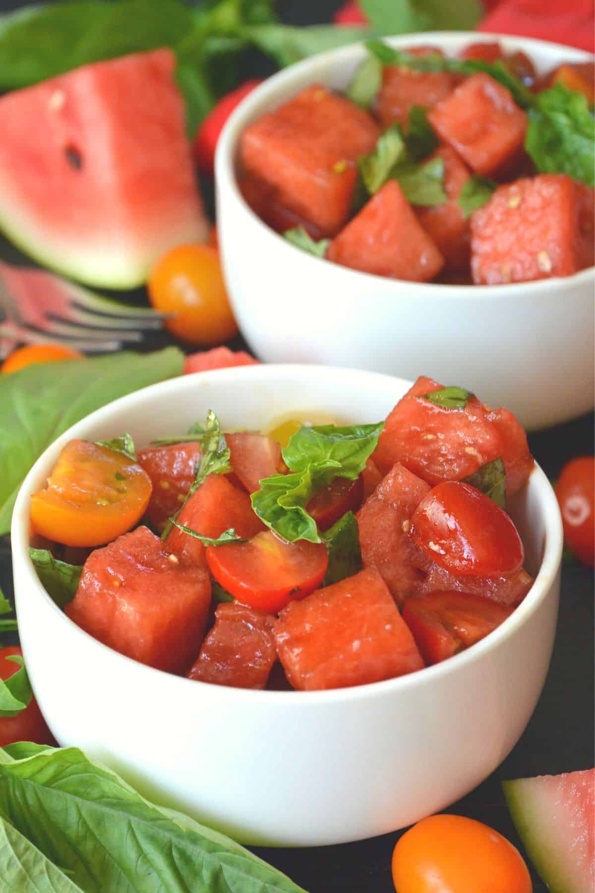 Tazones de ensalada de tomate y sandía adornados con albahaca fresca con una rodaja de melón en el fondo
