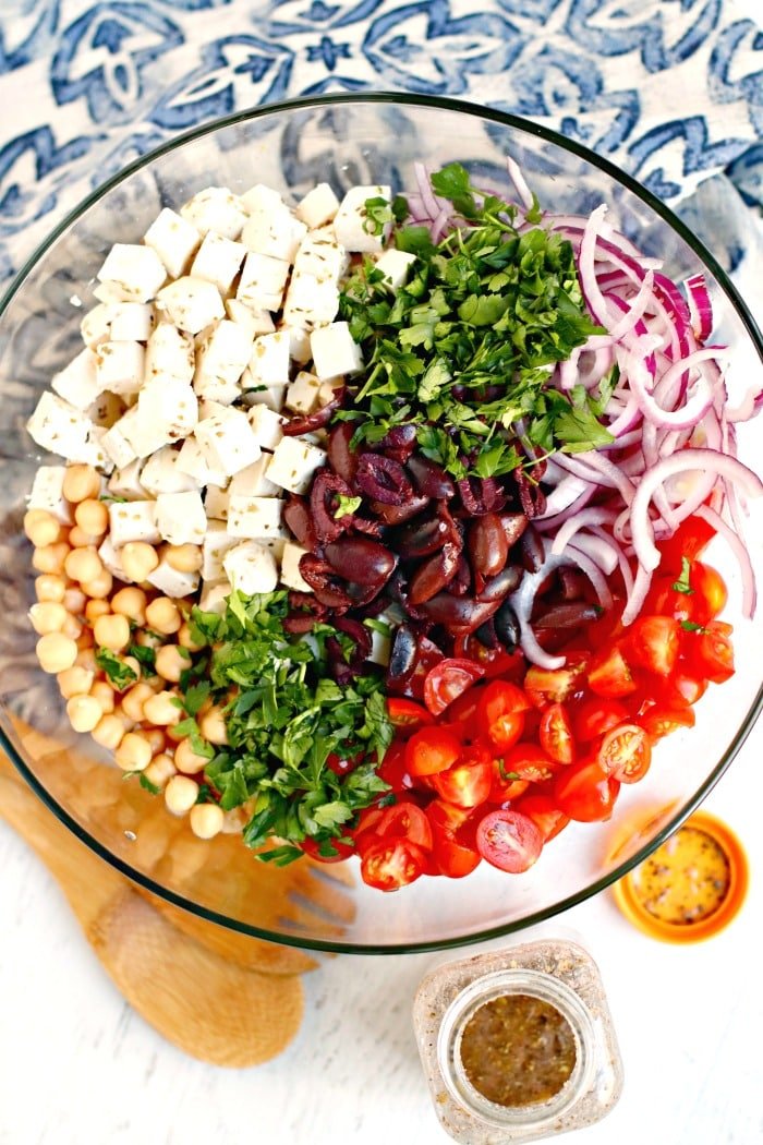 Ensaladera llena de los ingredientes para preparar una ensalada Orzo mediterránea y un tarro con ingredientes para el aderezo a un lado