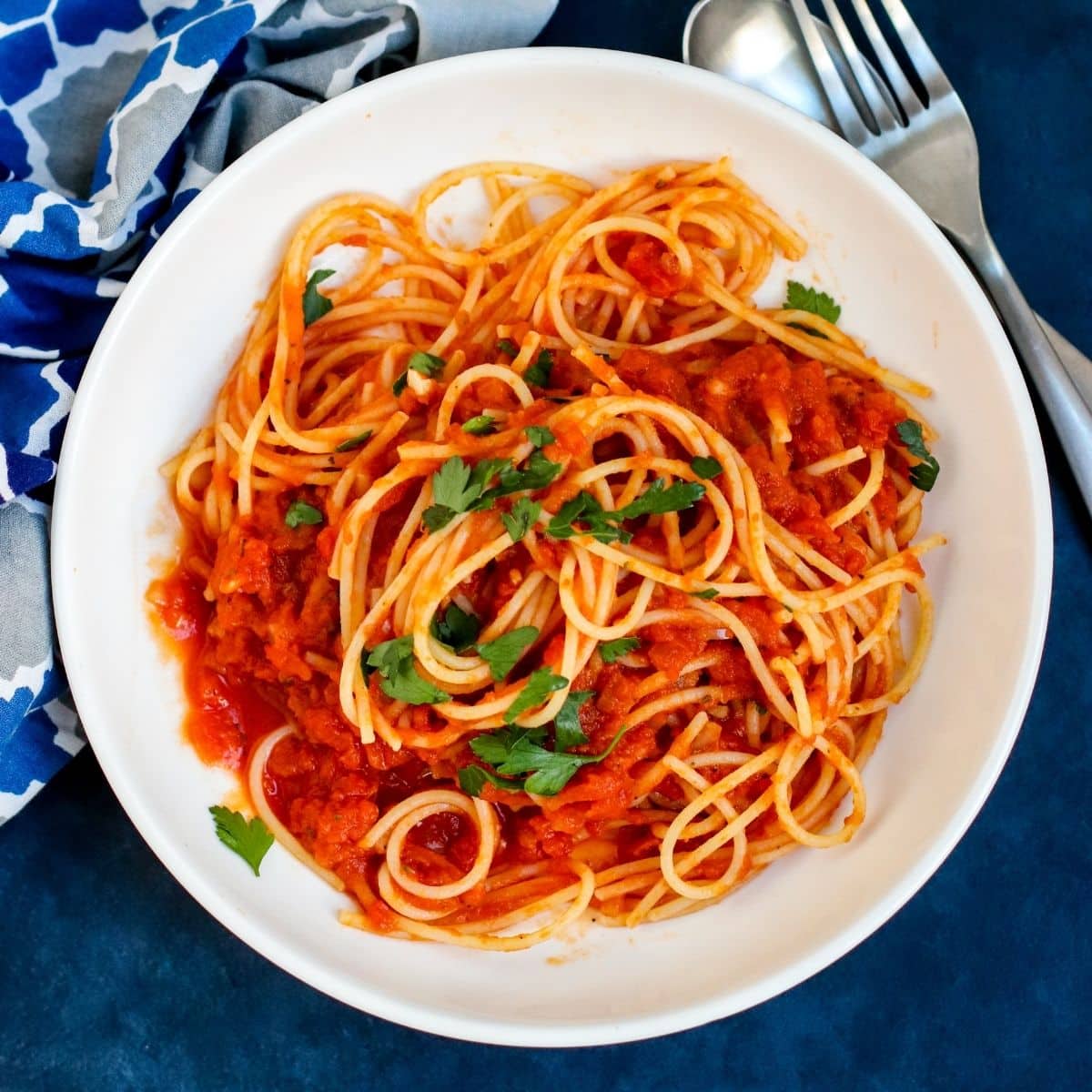 Cuenco de espagueti con calabaza marinara adornado con perejil fresco junto a un tenedor y una servilleta azul estampada.